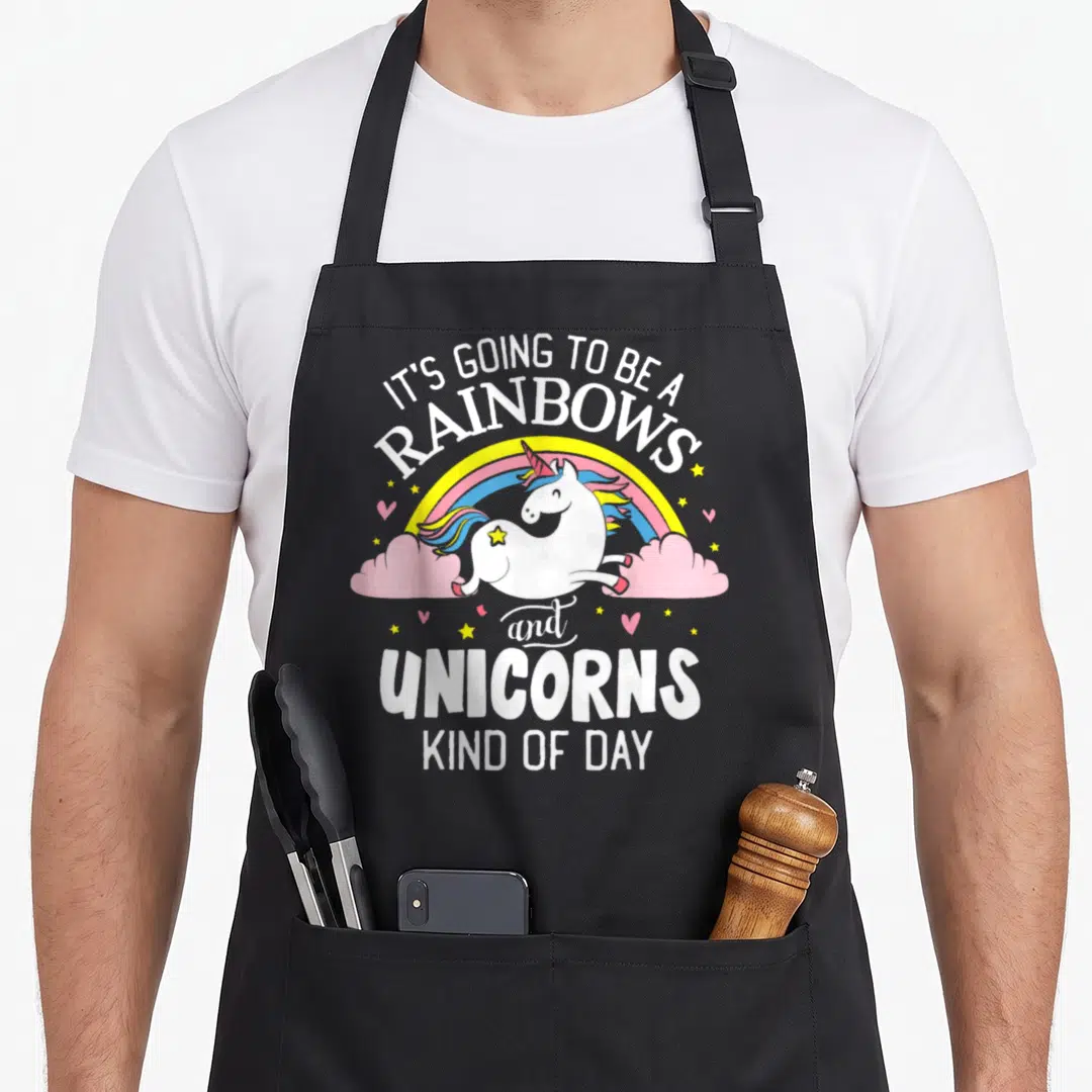 Unicorn rainbow apron - Cartoon unicorn jumping between pink clouds under arched text reading IT'S GOING TO BE — shown on a black cotton canvas apron against a white background
