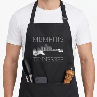 Souvenir memphis apron - White MEMPHIS TENNESSEE text surrounding a guitar silhouette filled with a city skyline — shown on a black cotton canvas apron against a white background