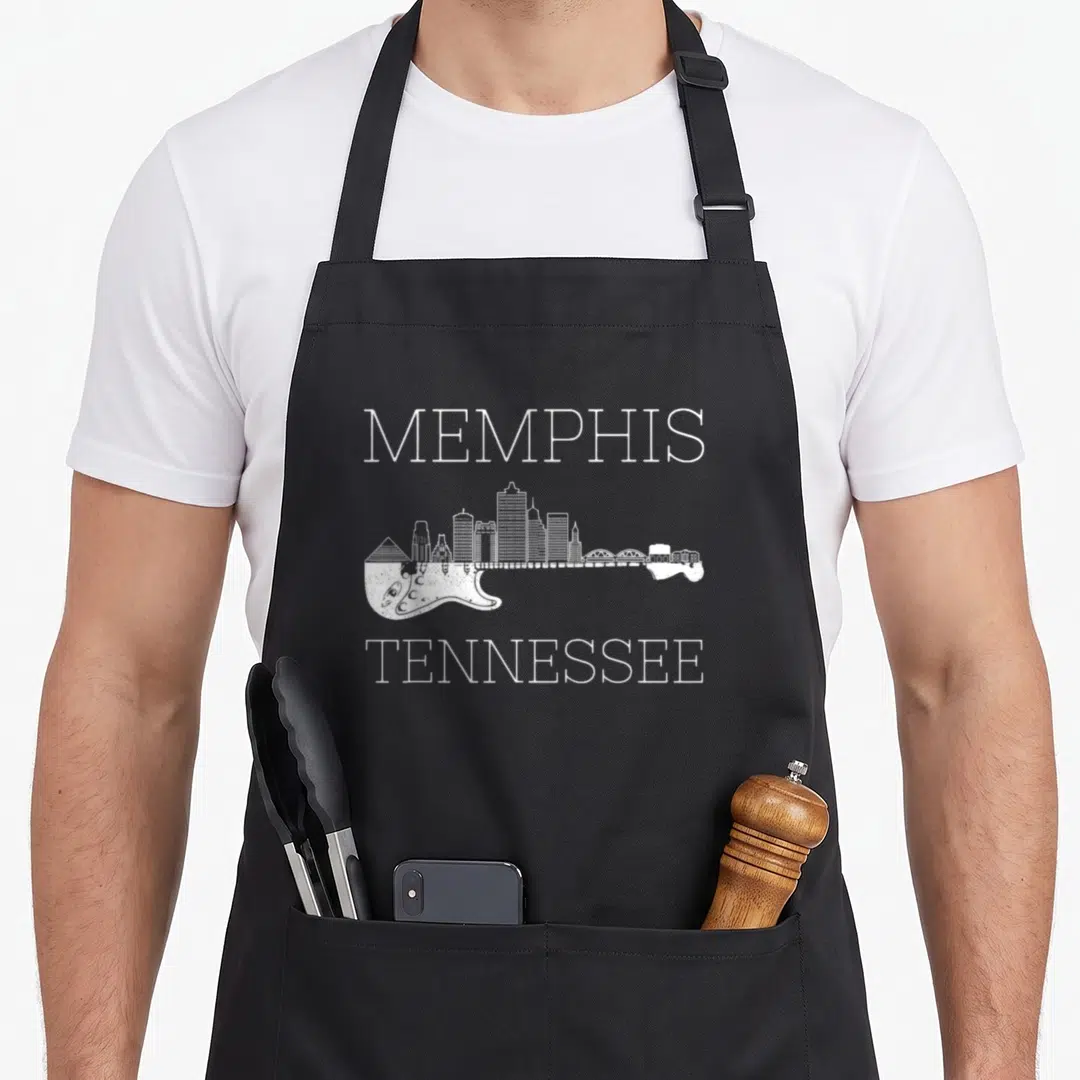 Souvenir memphis apron - White MEMPHIS TENNESSEE text surrounding a guitar silhouette filled with a city skyline — shown on a black cotton canvas apron against a white background