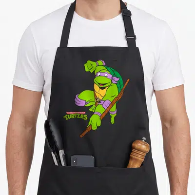 Donatello teenage mutant ninja apron - Green cartoon turtle with purple mask holding brown staff next to TEENAGE MUTANT — shown on a black cotton canvas apron against a white background