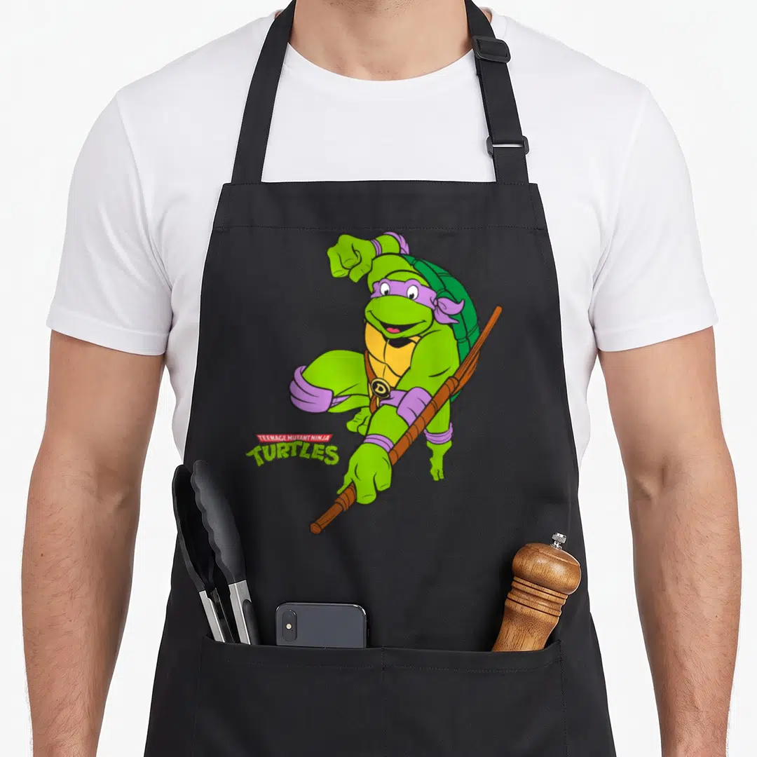Donatello teenage mutant ninja apron - Green cartoon turtle with purple mask holding brown staff next to TEENAGE MUTANT — shown on a black cotton canvas apron against a white background