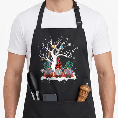 Gnome buffalo plaid christmas apron - Three gnomes in plaid hats sitting under a white tree with colorful lights — shown on a black cotton canvas apron against a white background