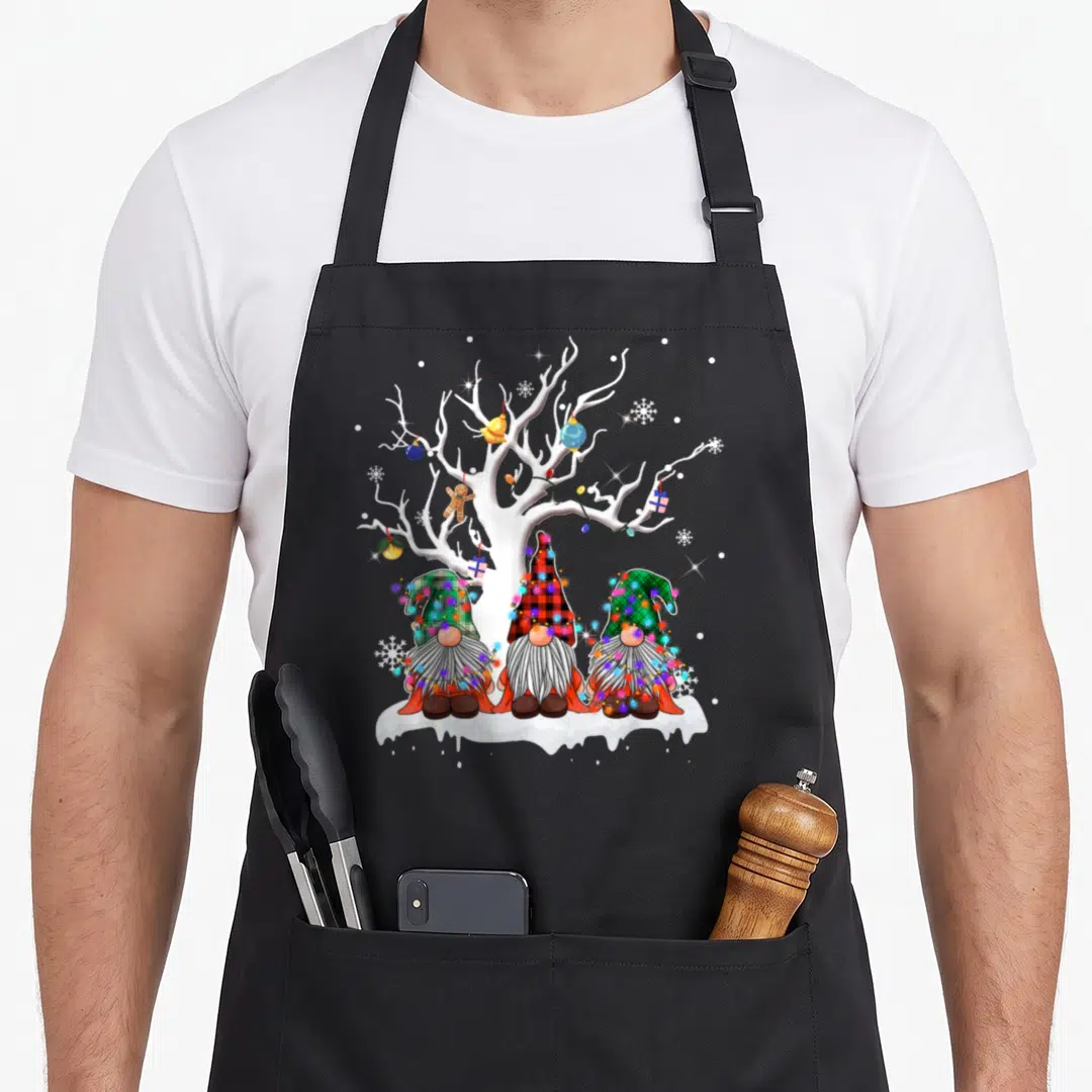Gnome buffalo plaid christmas apron - Three gnomes in plaid hats sitting under a white tree with colorful lights — shown on a black cotton canvas apron against a white background