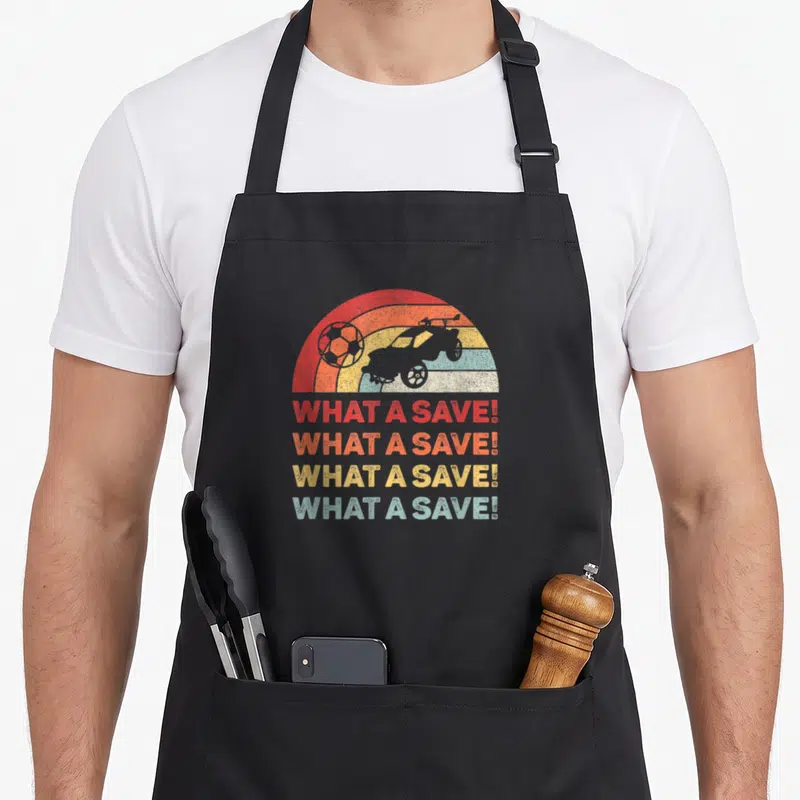 Save car apron - Retro car and soccer ball over rainbow arc with four stacked What A Save texts in red orange yellow — shown on a black cotton canvas apron against a white background