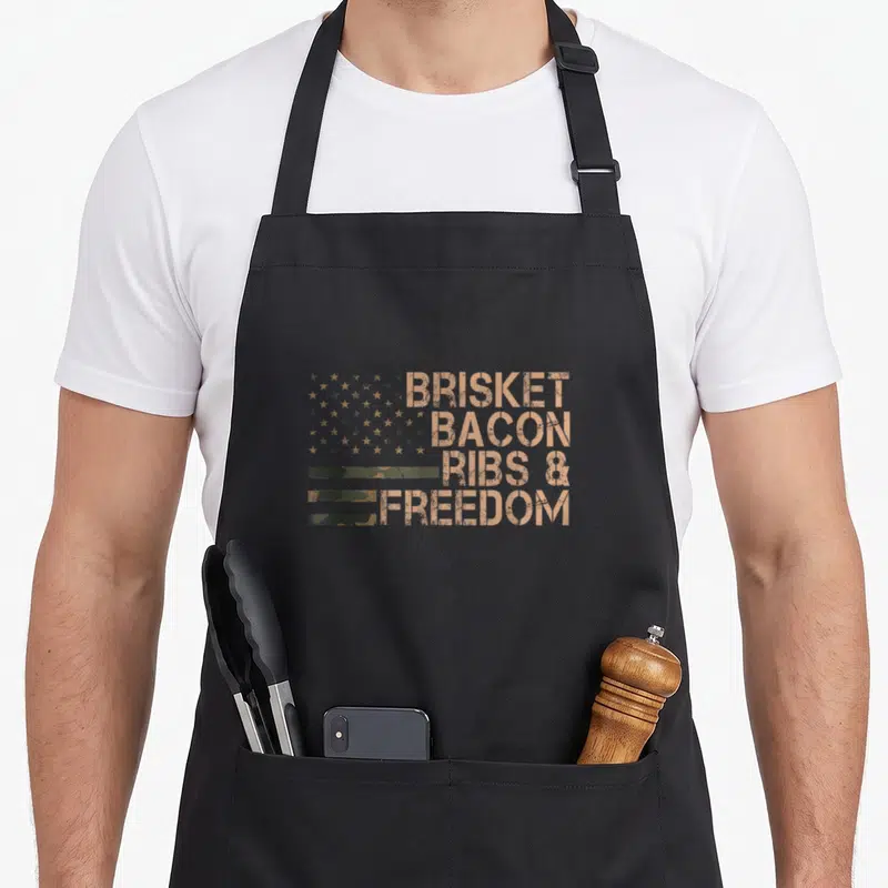 Brisket meat smoking apron - Distressed tan text BRISKET BACON RIBS FREEDOM with olive star cluster and stripes — shown on a black cotton canvas apron against a white background