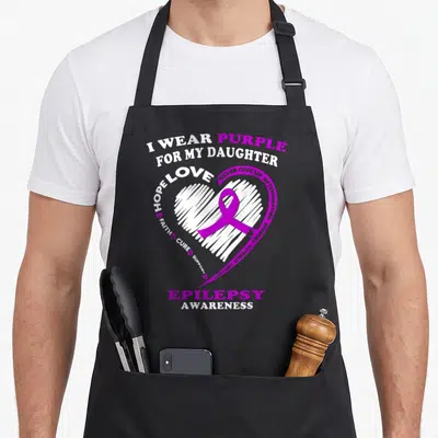 Daughter epilepsy awareness apron - White scribbled heart with purple ribbon and epilepsy awareness text — shown on a black cotton canvas apron against a white background