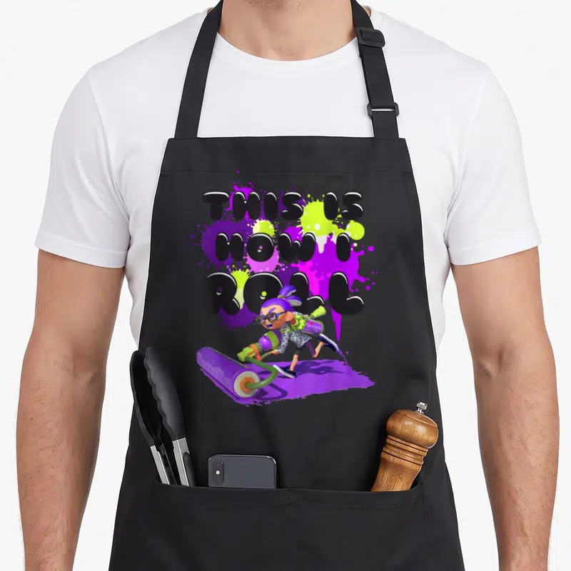 Inkling roll apron - Cartoon Inkling pushing purple roller under THIS IS HOW I ROLL text over purple and lime splatters — shown on a black cotton canvas apron against a white background