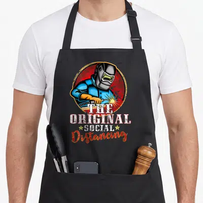Vintage welding welder dad apron - Welder in blue helmet holding torch below text The Original Social Distancing — shown on a black cotton canvas apron against a white background