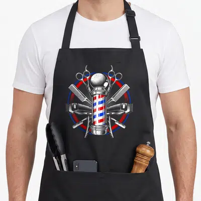 Barber hairstylist apron - Barber pole with scissors combs clippers razors in red white blue circle — shown on a black cotton canvas apron against a white background