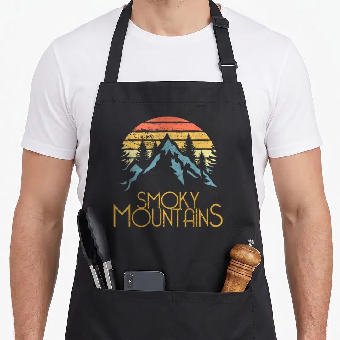 Smoky lookout mountain apron - Retro graphic of blue mountains and pine trees behind a striped sun with stacked SMOKY — shown on a black cotton canvas apron against a white background