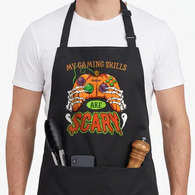 Skill halloween apron - Pumpkin controller held by skeleton hands with text My Gaming Skills Are Scary flanked by two — shown on a black cotton canvas apron against a white background