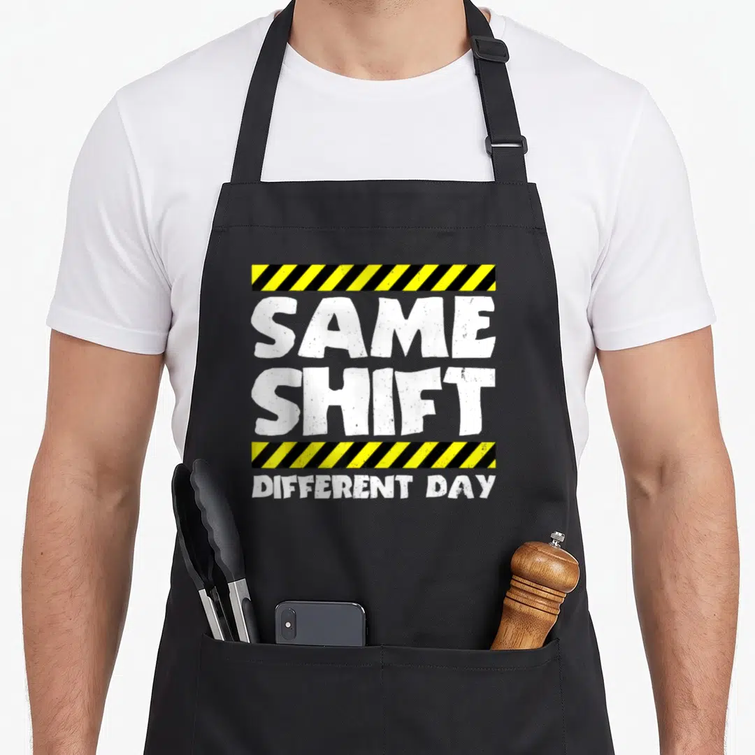 Shift meme apron - White same shift different day text between yellow and black striped bars — shown on a black cotton canvas apron against a white background
