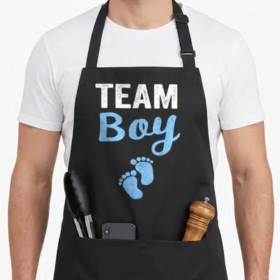 Gender reveal shower apron - Team Boy text with two blue distressed baby footprints on black fabric — shown on a black cotton canvas apron against a white background