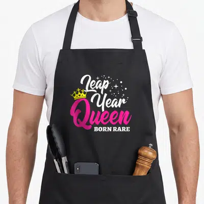 Rare birthday apron - Pink Queen text with yellow crown and white stars reading Leap Year Born Rare — shown on a black cotton canvas apron against a white background