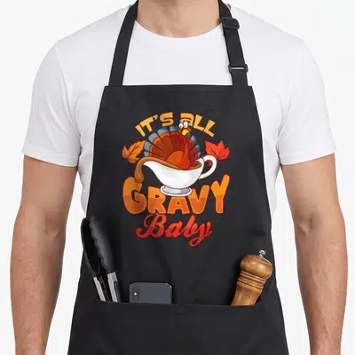 Gravy thanksgiving apron - Cartoon turkey in white gravy boat with two autumn leaves and IT'S ALL GRAVY Baby text — shown on a black cotton canvas apron against a white background
