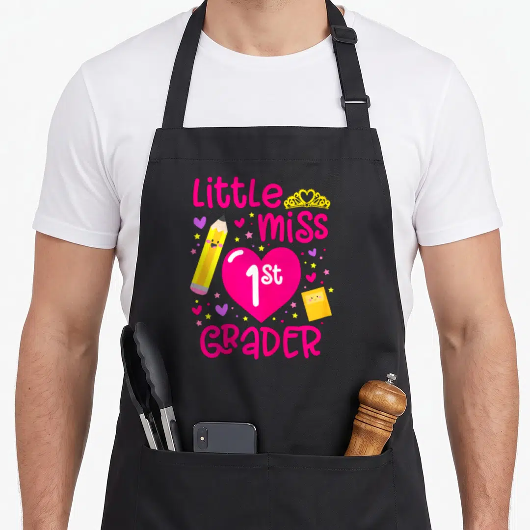 1st miss grade apron - Little miss 1st GRADER text with pink heart, yellow pencil, and notebook — shown on a black cotton canvas apron against a white background