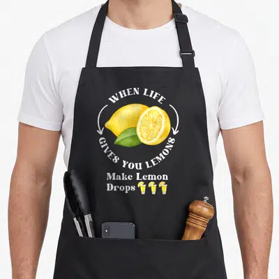 Give drop for love and lemons apron - watercolor lemons and three shot glasses with circular white text — shown on a black cotton canvas apron against a white background