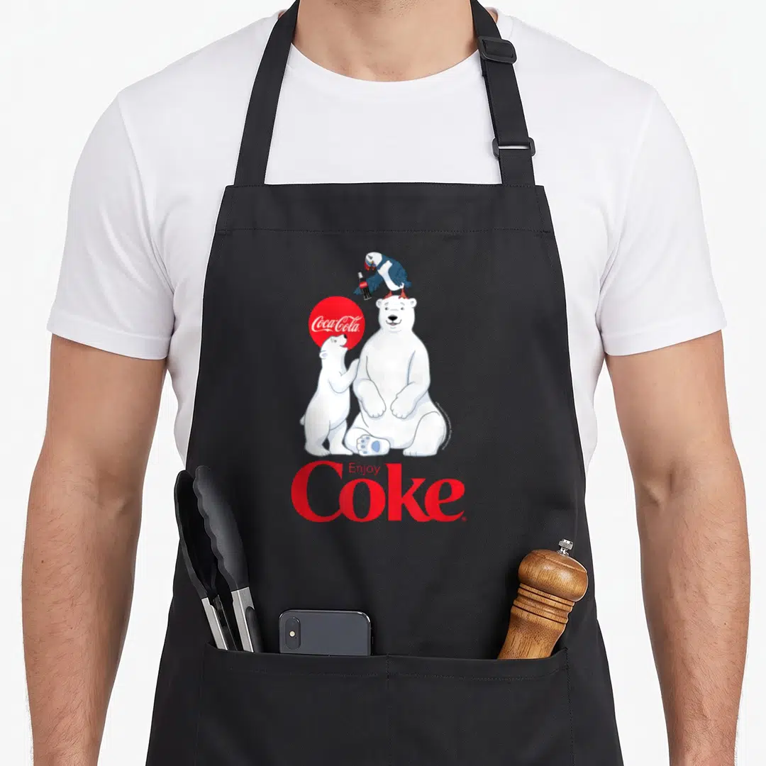 Cola bear apron - Cartoon polar bear and cub with parrot holding bottle, red Coca-Cola circle and Enjoy Coke text — shown on a black cotton canvas apron against a white background