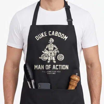 Caboom dog breed aprons - White cartoon Duke Caboom toy standing in front of motorcycle with arched text — shown on a black cotton canvas apron against a white background