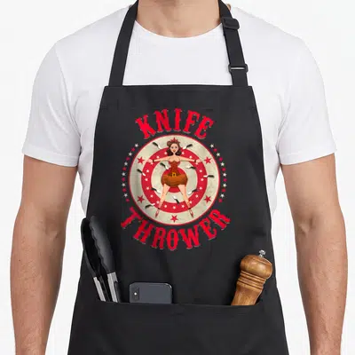 Circus knife apron - Cartoon woman on target with knives and KNIFE THROWER text in red and brown — shown on a black cotton canvas apron against a white background