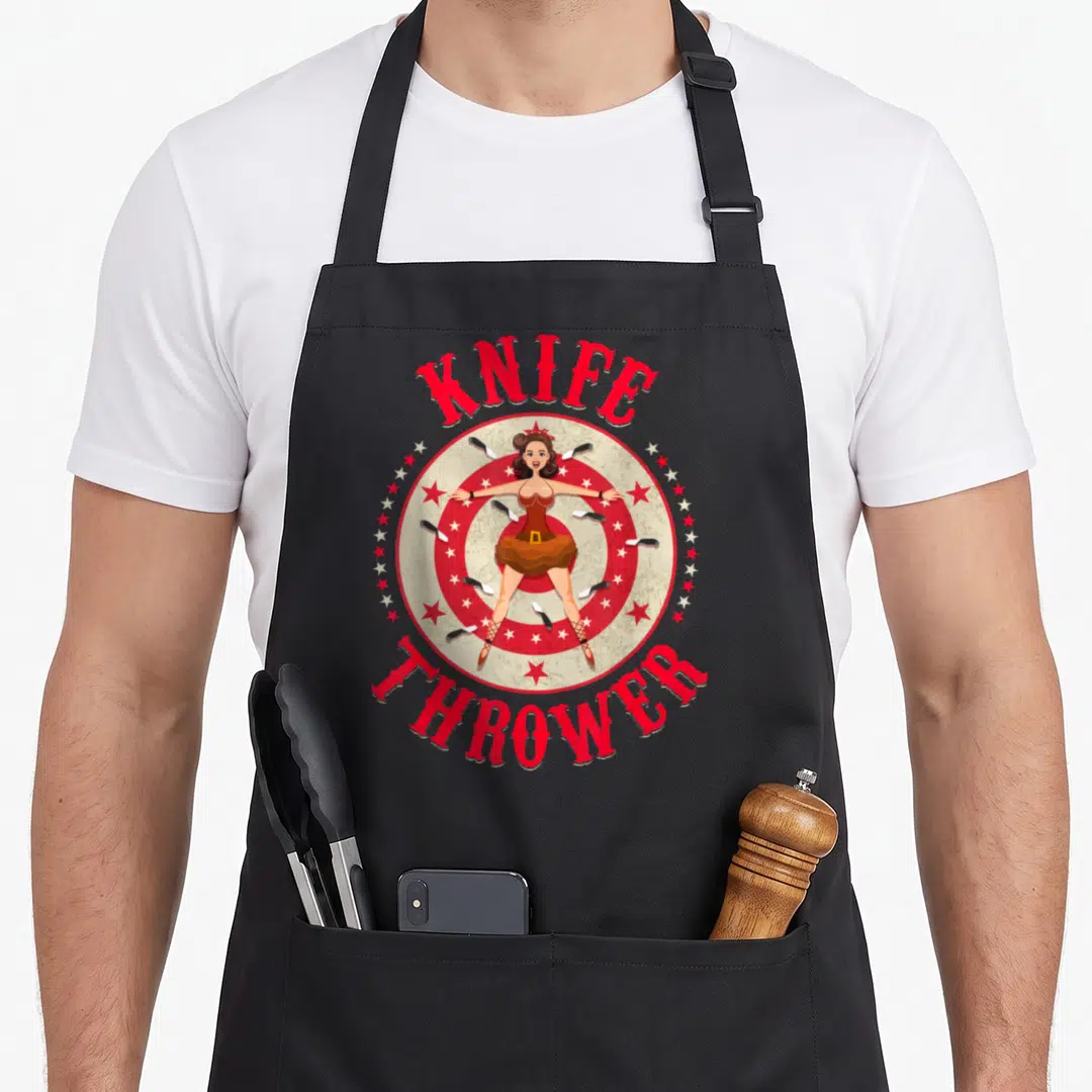 Circus knife apron - Cartoon woman on target with knives and KNIFE THROWER text in red and brown — shown on a black cotton canvas apron against a white background