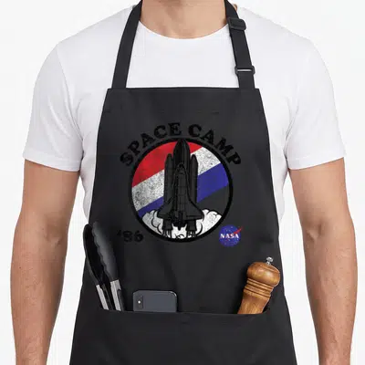 Camp nasa apron - Space shuttle inside red white blue circle with SPACE CAMP '86 text and distressed texture — shown on a black cotton canvas apron against a white background