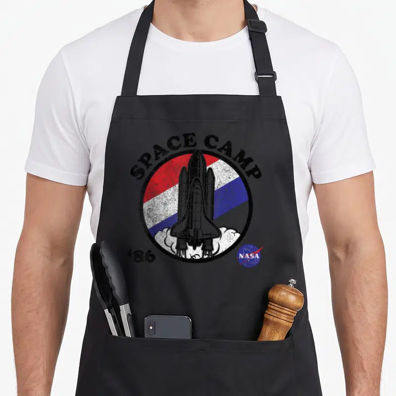 Camp nasa apron - Space shuttle inside red white blue circle with SPACE CAMP '86 text and distressed texture — shown on a black cotton canvas apron against a white background