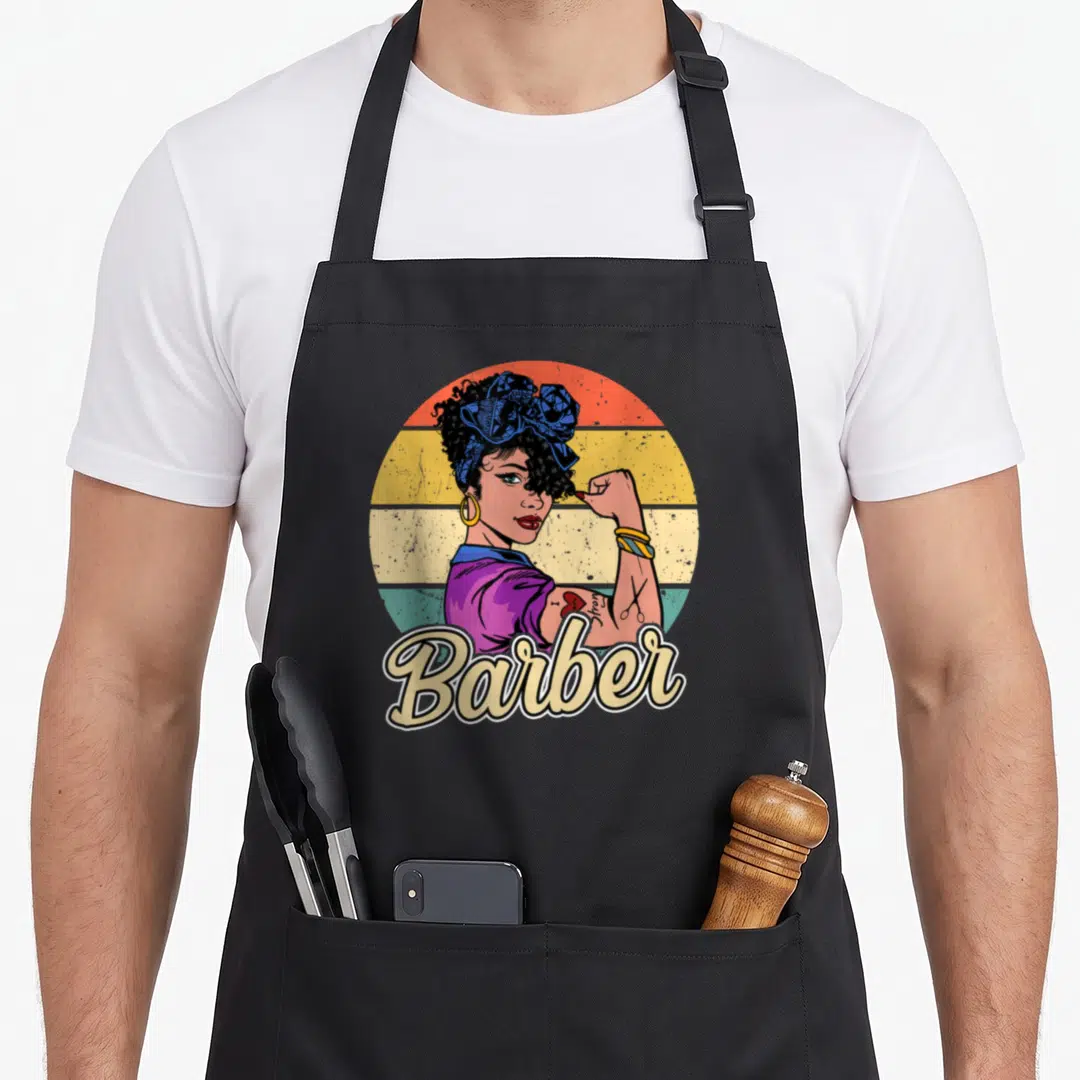 Funny barber apron - Vintage woman flexing arm with scissors tattoo and Barber script on striped background — shown on a black cotton canvas apron against a white background