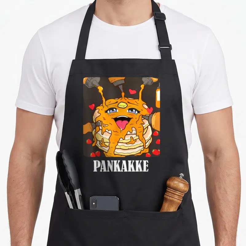 Pun funny naughty aprons - Cartoon pancake stack with face, syrup, strawberries, hearts, and PANKAKKE text — shown on a black cotton canvas apron against a white background