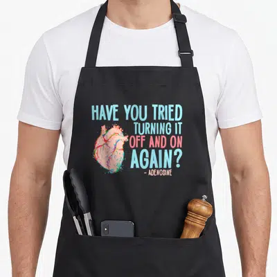 Adenosine i turn grills on apron - Anatomical heart watercolor with text Have You Tried Turning It Off And On Again — shown on a black cotton canvas apron against a white background