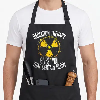 Certain radiation aprons - Yellow radiation symbol with white text RADIATION THERAPY GIVES YOU THAT CERTAIN GLOW — shown on a black cotton canvas apron against a white background