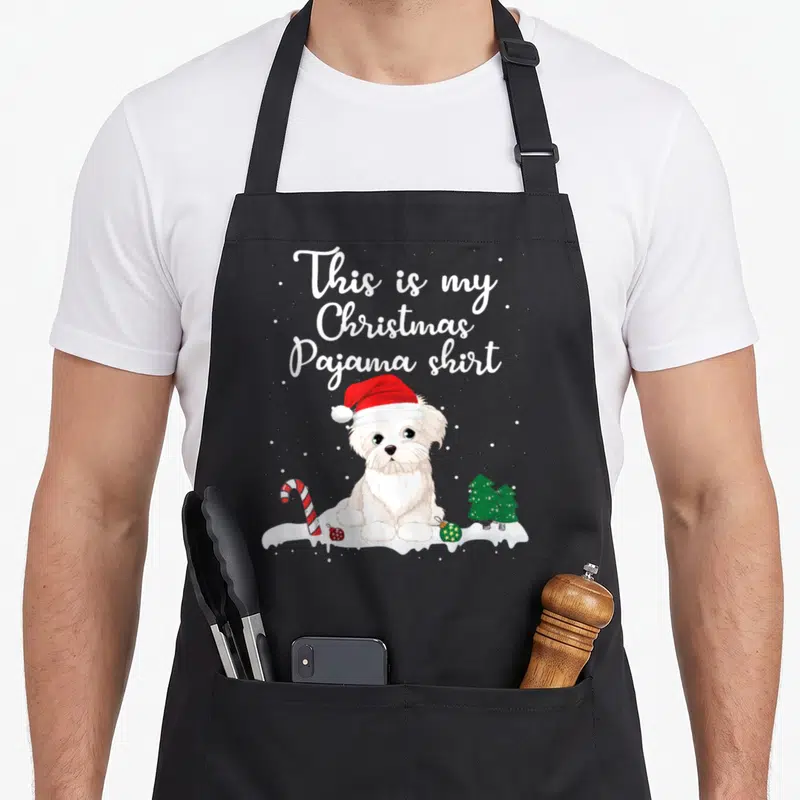 Pajama christmas dog apron - Cartoon white dog in red hat with This is my Christmas Pajama shirt text and candy cane — shown on a black cotton canvas apron against a white background