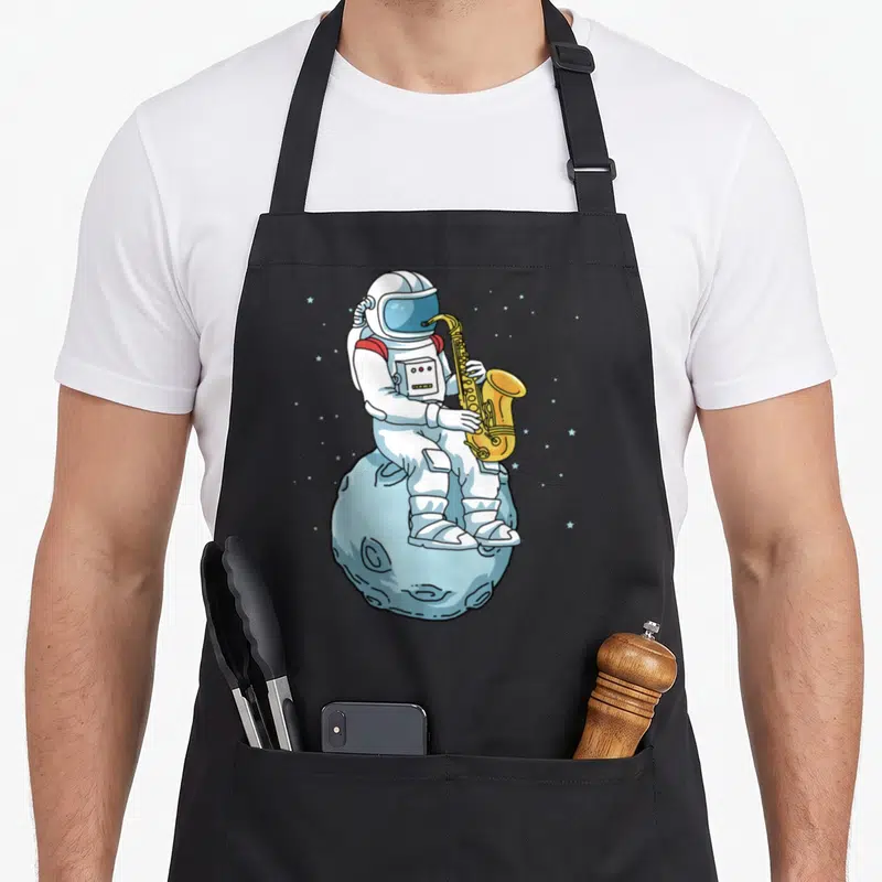 Saxophone saxophonist apron - Cartoon astronaut playing yellow saxophone sitting on light blue moon with stars — shown on a black cotton canvas apron against a white background