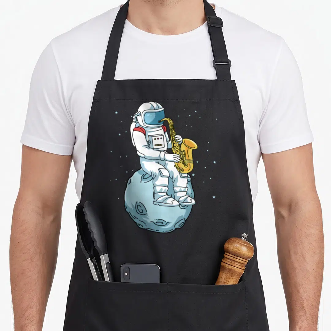 Saxophone saxophonist apron - Cartoon astronaut playing yellow saxophone sitting on light blue moon with stars — shown on a black cotton canvas apron against a white background