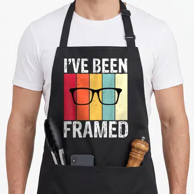 Framed optometrist apron - I've Been Framed text with black glasses over five vertical colored stripes — shown on a black cotton canvas apron against a white background