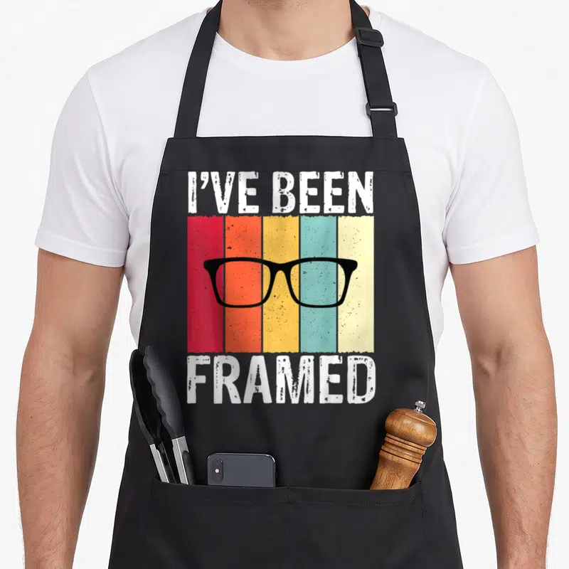 Framed optometrist apron - I've Been Framed text with black glasses over five vertical colored stripes — shown on a black cotton canvas apron against a white background