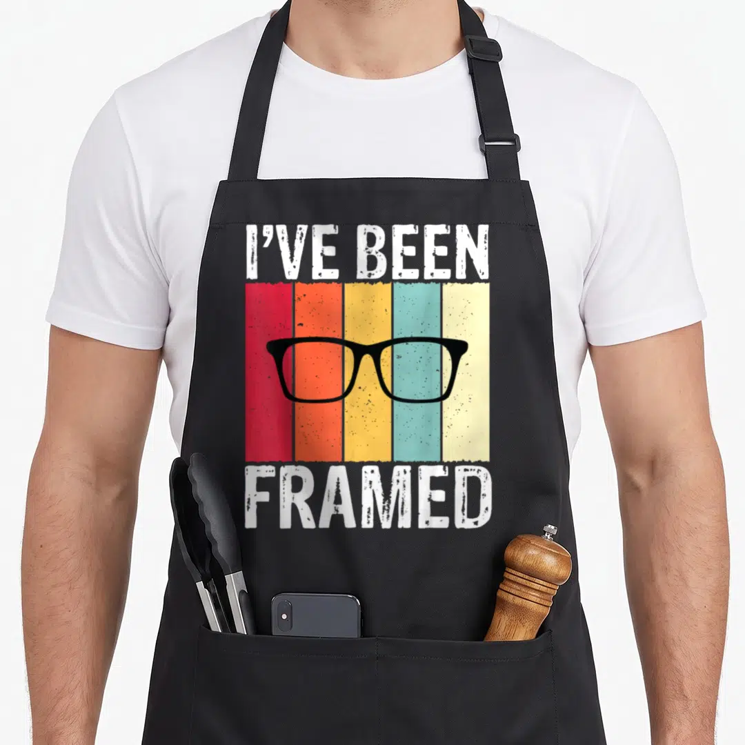 Framed optometrist apron - I've Been Framed text with black glasses over five vertical colored stripes — shown on a black cotton canvas apron against a white background