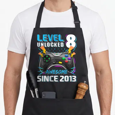 8th birthday video game apron - Black game controller with colorful buttons surrounded by Level 8 Unlocked text — shown on a black cotton canvas apron against a white background