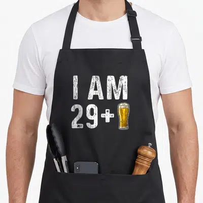 Drinker birthday apron - I AM 29 plus yellow beer glass illustration in distressed white font — shown on a black cotton canvas apron against a white background