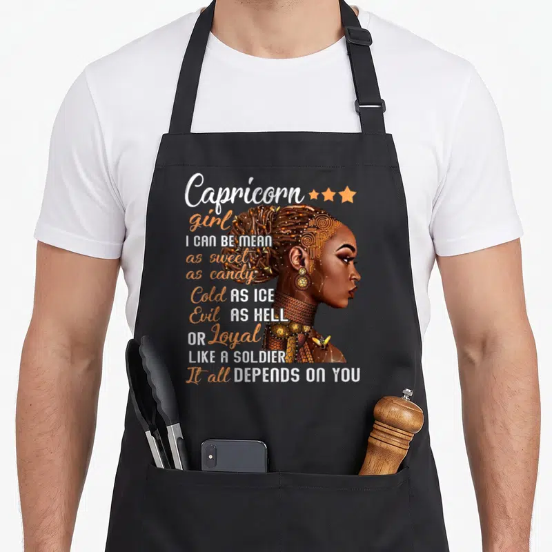 December january birthday apron - Capricorn girl illustration with stacked text about being mean, sweet, cold, evil — shown on a black cotton canvas apron against a white background