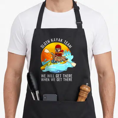 Kayaking sloth apron - Cartoon sloth in yellow kayak with red helmet and SLOTH KAYAK TEAM text against orange sun — shown on a black cotton canvas apron against a white background