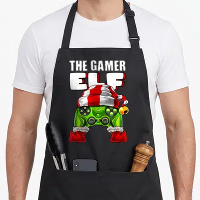 Family video christmas elf apron - Green game controller wearing a red and white striped elf hat and red boots — shown on a black cotton canvas apron against a white background