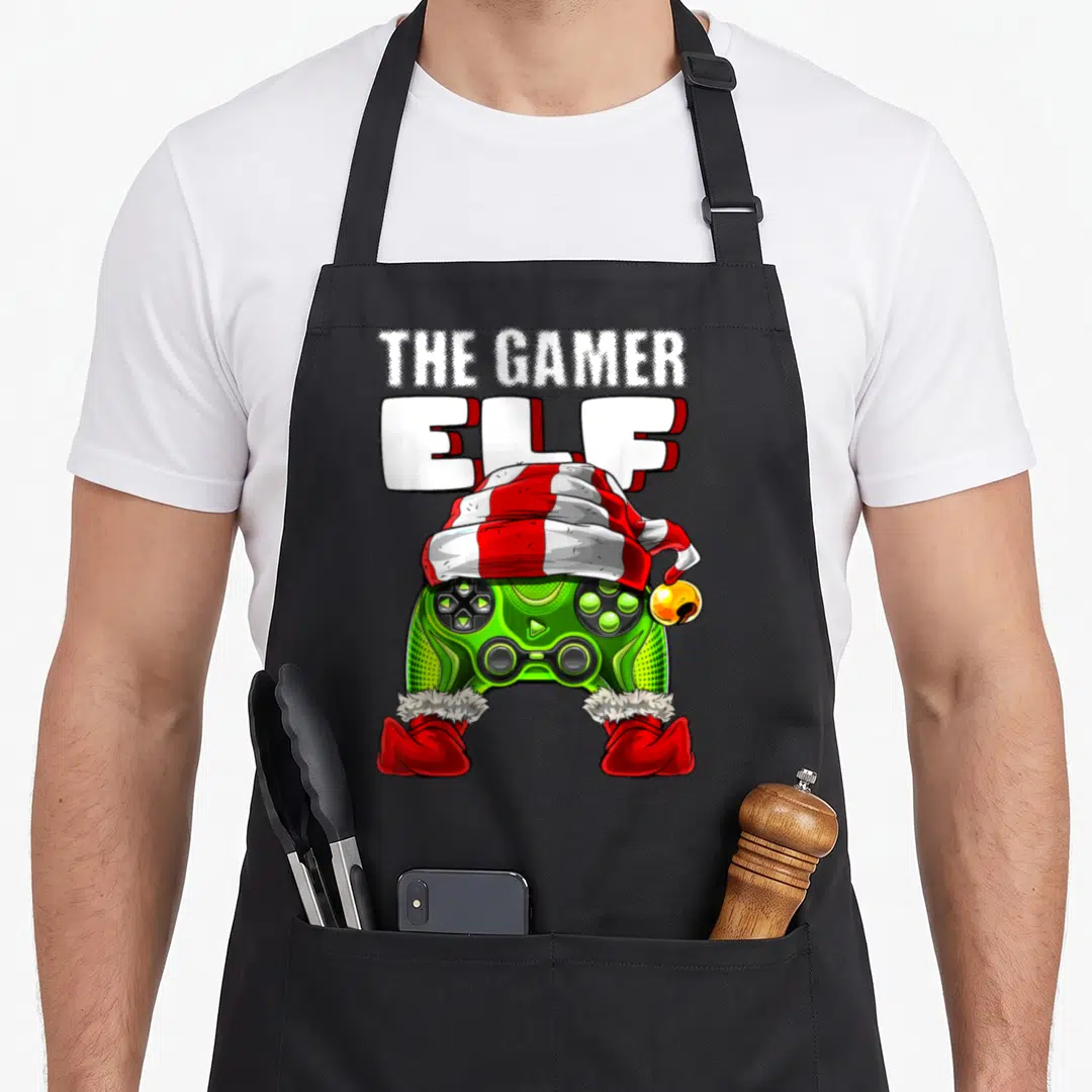 Family video christmas elf apron - Green game controller wearing a red and white striped elf hat and red boots — shown on a black cotton canvas apron against a white background