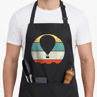 Cute hot balloon apron - Grey hot air balloon over yellow teal white orange stripes with mountain base — shown on a black cotton canvas apron against a white background
