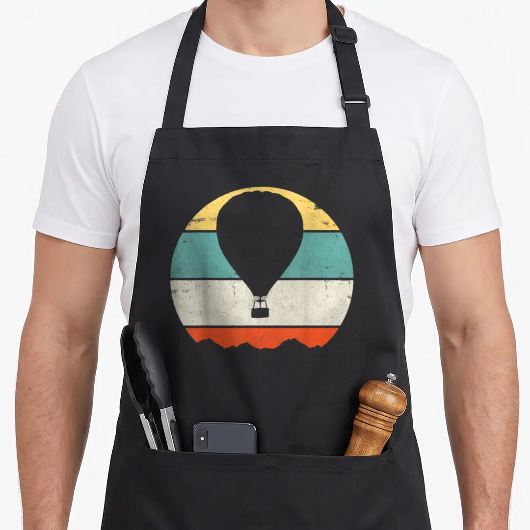 Cute hot balloon apron - Grey hot air balloon over yellow teal white orange stripes with mountain base — shown on a black cotton canvas apron against a white background
