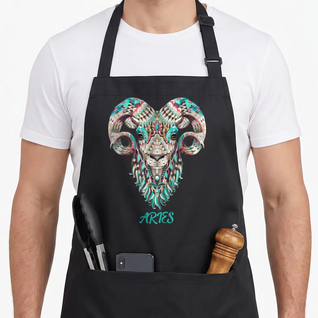 Ary birthday apron - Ornate Aries ram head in teal beige pink line art with text below — shown on a black cotton canvas apron against a white background