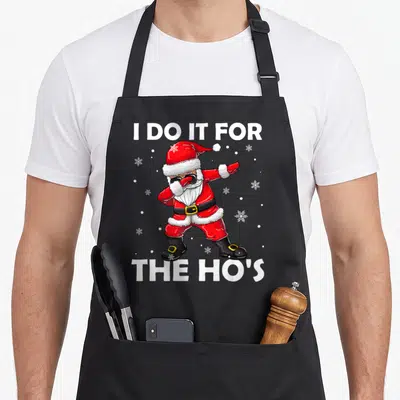 Hos santa claus apron - Cartoon Santa dabbing between text I DO IT FOR THE HO'S with snowflakes — shown on a black cotton canvas apron against a white background