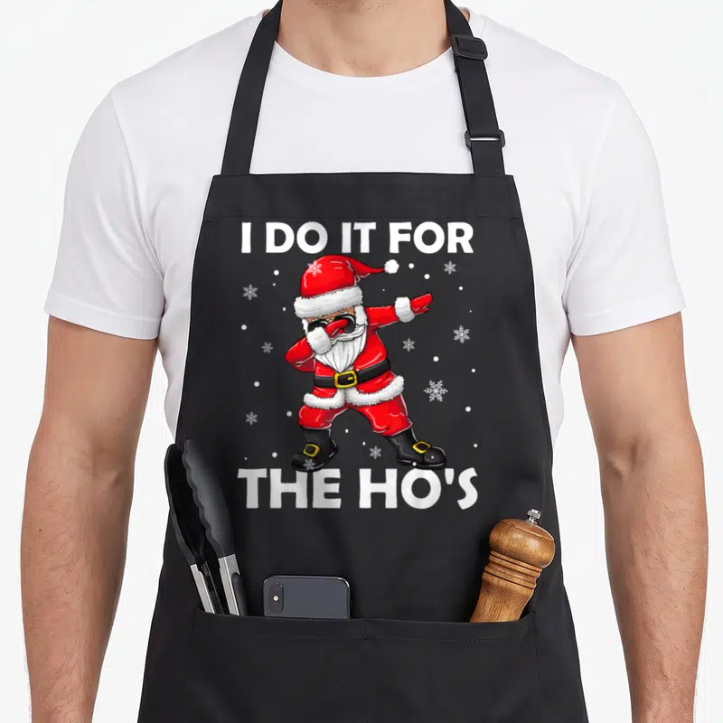 Hos santa claus apron - Cartoon Santa dabbing between text I DO IT FOR THE HO'S with snowflakes — shown on a black cotton canvas apron against a white background