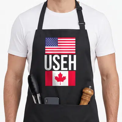 Useh pride american flag apron - USA flag above USEH text and Canada flag below in red white blue — shown on a black cotton canvas apron against a white background