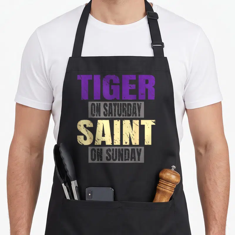 Tiger funny sunday apron - Stacked purple and yellow distressed text reading Tiger On Saturday Saint On Sunday — shown on a black cotton canvas apron against a white background